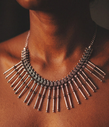 A close-up of a model wearing a silver-toned statement necklace with multiple vertical bar elements and a textured chain, shown against warm skin tones.