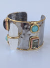 A hammered silver-tone cuff bracelet with gold trim, featuring a large clear crystal, a round turquoise stone, a rectangular green gemstone, and a dark oval stone.