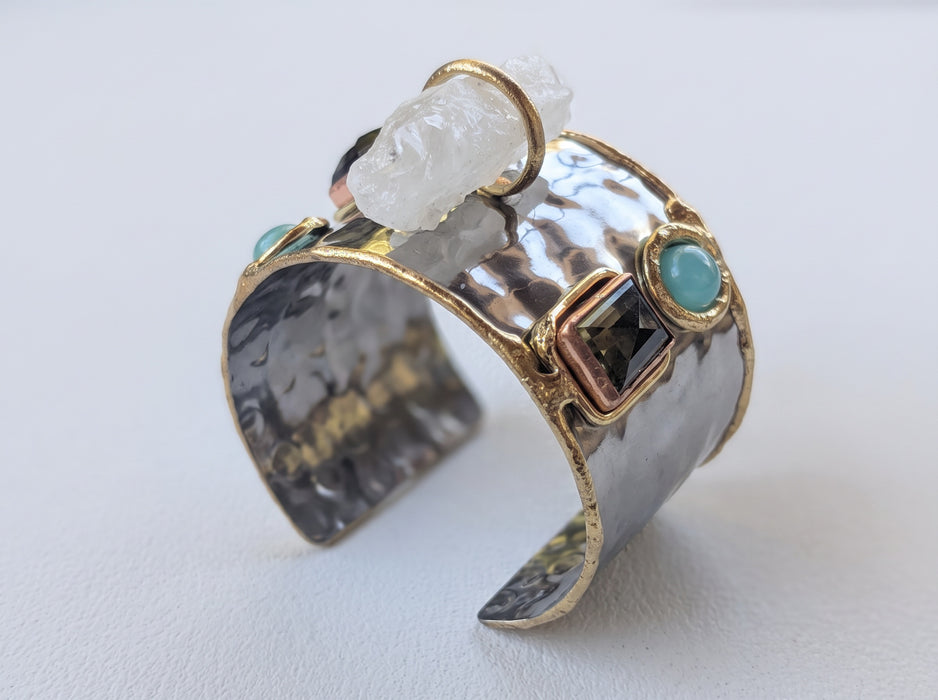 A hammered silver cuff ring with gold trim, turquoise-colored stones, a dark square stone, and a chunky white crystal on a light gray background.
