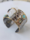 A hammered silver-tone cuff ring with gold-tone trim, turquoise stones, a clear quartz crystal, and a square iridescent gemstone, displayed on a light surface.