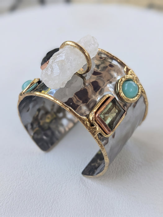 A hammered silver-tone cuff ring with gold-tone trim, turquoise stones, a clear quartz crystal, and a square iridescent gemstone, displayed on a light surface.
