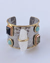 A hammered silver cuff bracelet with gold trim, featuring multiple inset gemstones in black, green, and turquoise, along with a central clear quartz crystal, set against a plain light gray background.