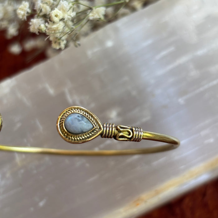 A gold-toned moonstone metal cuff bracelet with a teardrop-shaped light blue stone and wrapped wire detailing, displayed on a translucent white stone slab with dried flowers in the background.